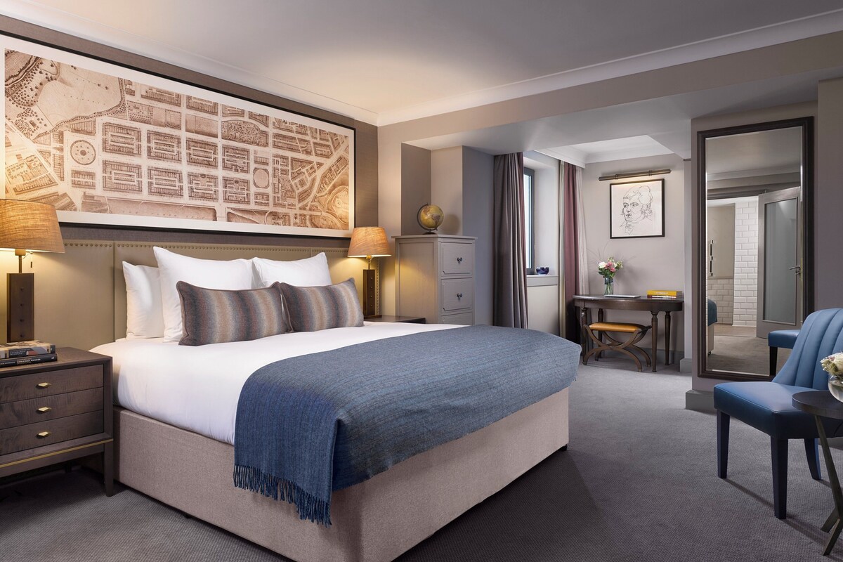 A hotel room with a large bed, a desk, a chair, a lamp, a globe, and a map on the wall.