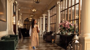 A hotel lobby with a checkered floor, green sofas, a woman in a beige coat, and a large window with a floral arrangement.