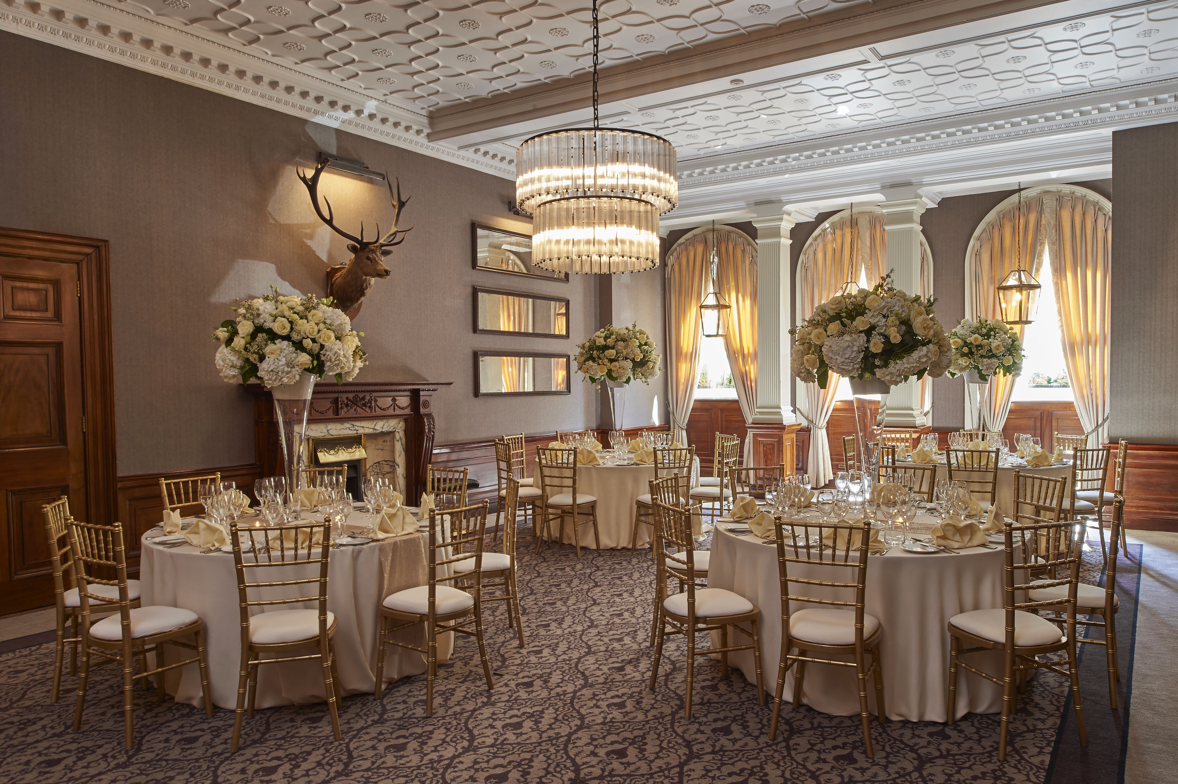 A banquet hall with round tables, gold chairs, chandeliers, and floral centerpieces.