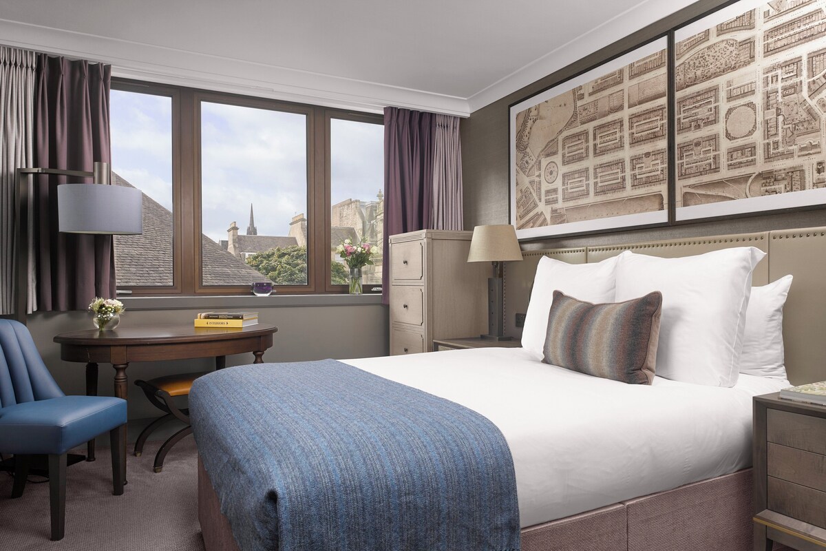 A hotel room with a large bed, a desk, a chair, a lamp, and a view of the cityscape through the window.