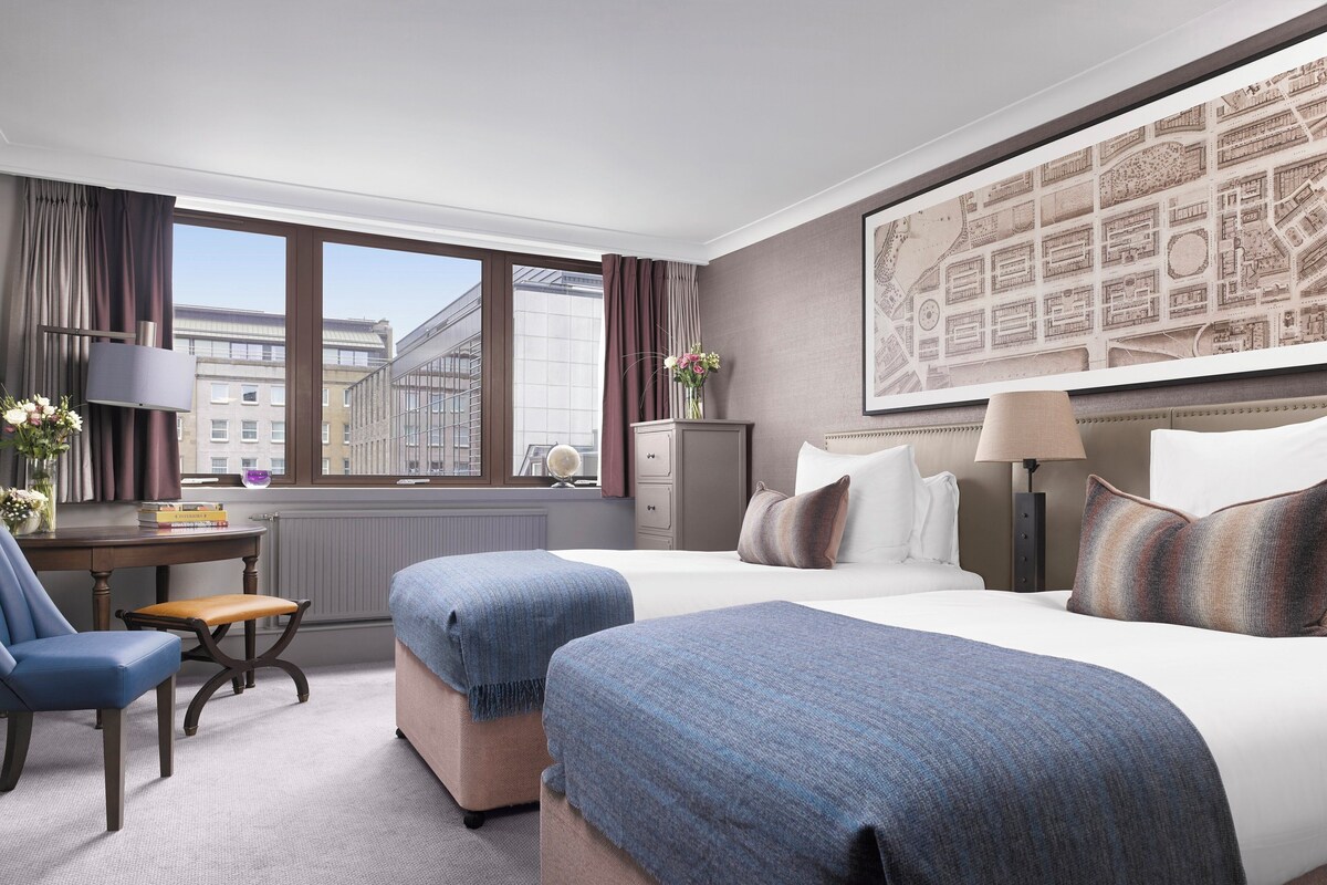 A hotel room with a large window, a desk with a lamp, two beds with blue and white bedding, a chair, and a map on the wall.