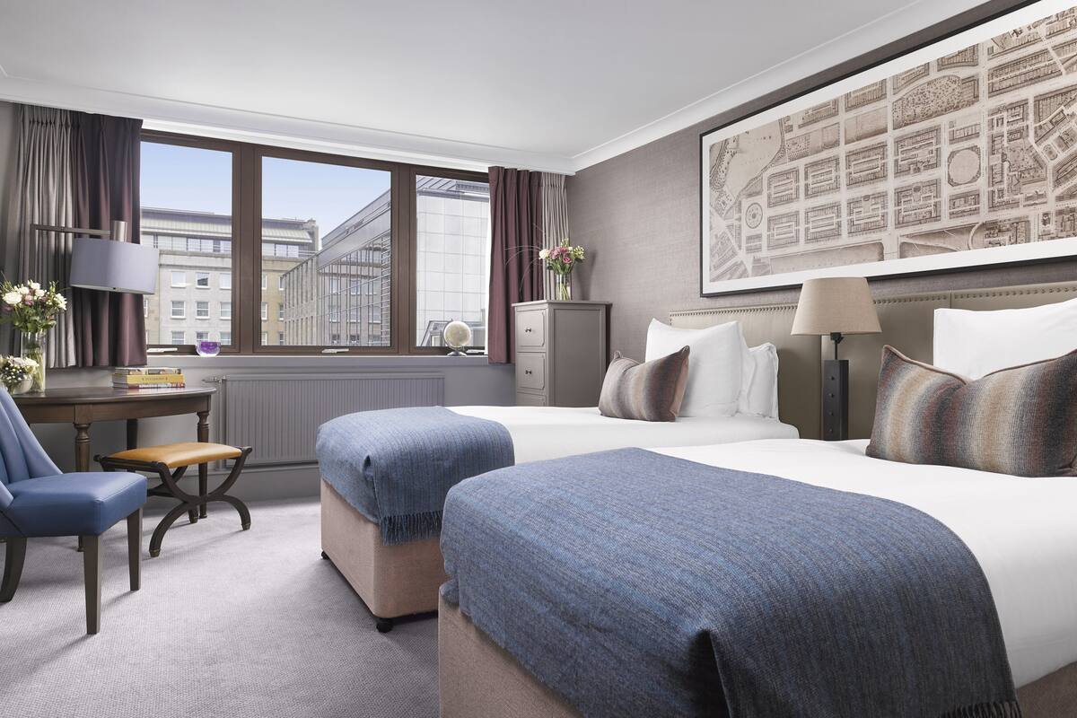 Et hotelværelse med et stort vindue, et skrivebord med lampe, to senge med blåt og hvidt sengetøj, en stol og et kort på væggen.