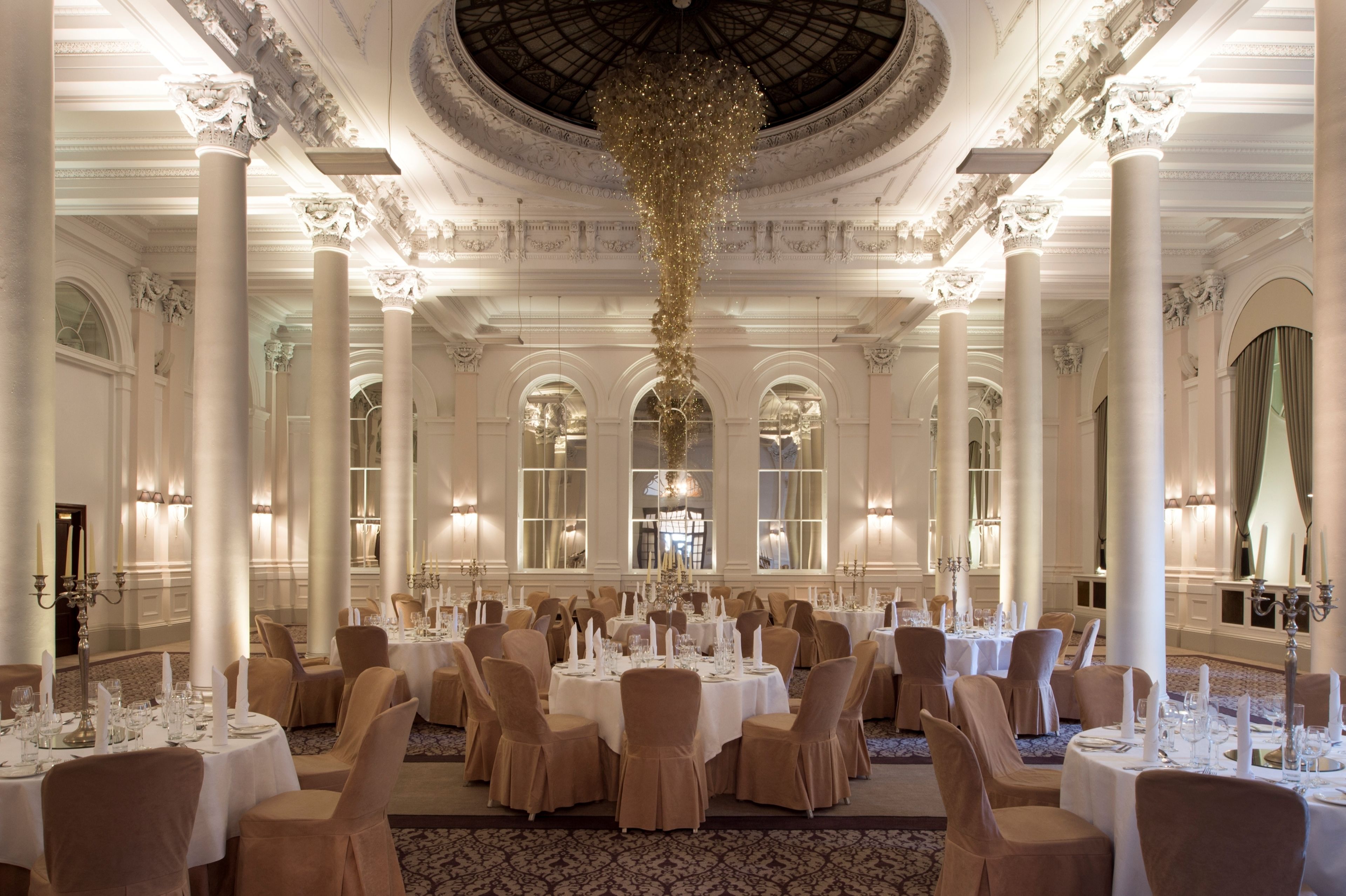 A grand ballroom with ornate columns, chandeliers, and a high ceiling. Tables are set for a formal event.