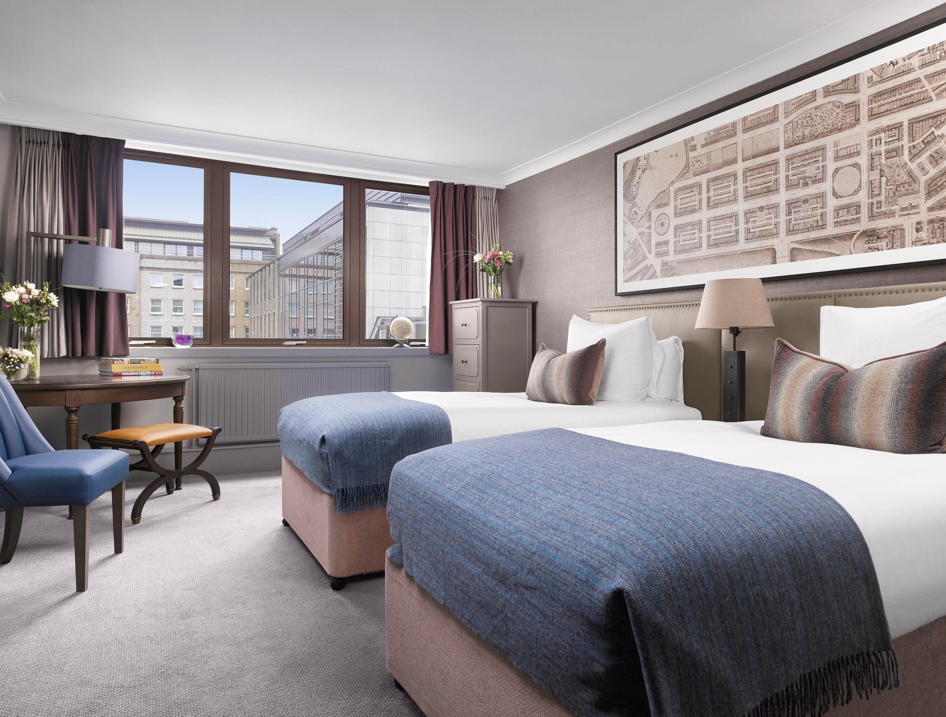 Ein Hotelzimmer mit zwei Betten, einem Schreibtisch, einem Stuhl, einer Lampe, einer Karte an der Wand und Blick auf Gebäude vor dem Fenster.