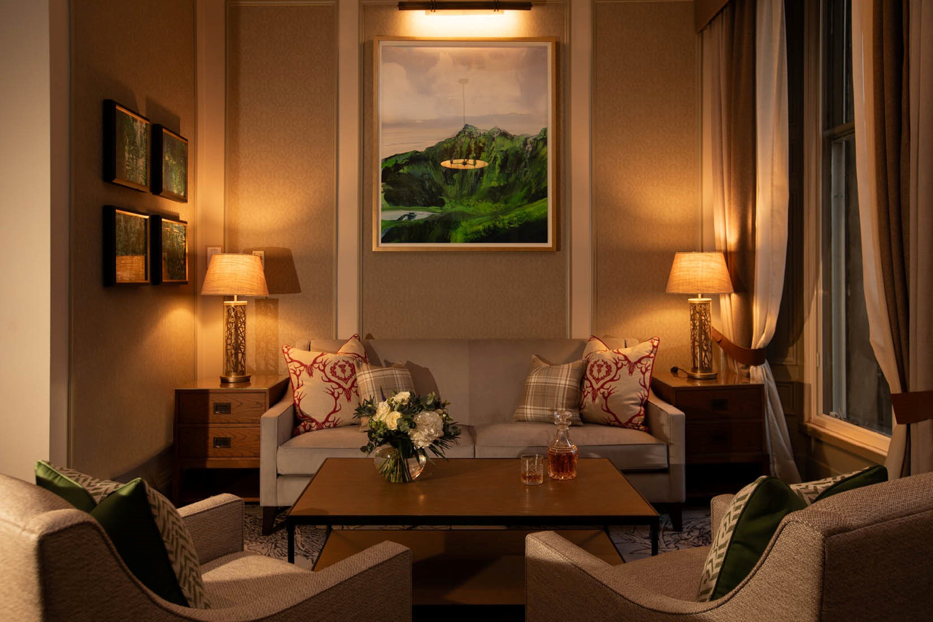 A well-lit living room with a sofa set, two side tables with lamps, a coffee table with a vase of flowers, and a framed landscape painting on the wall.