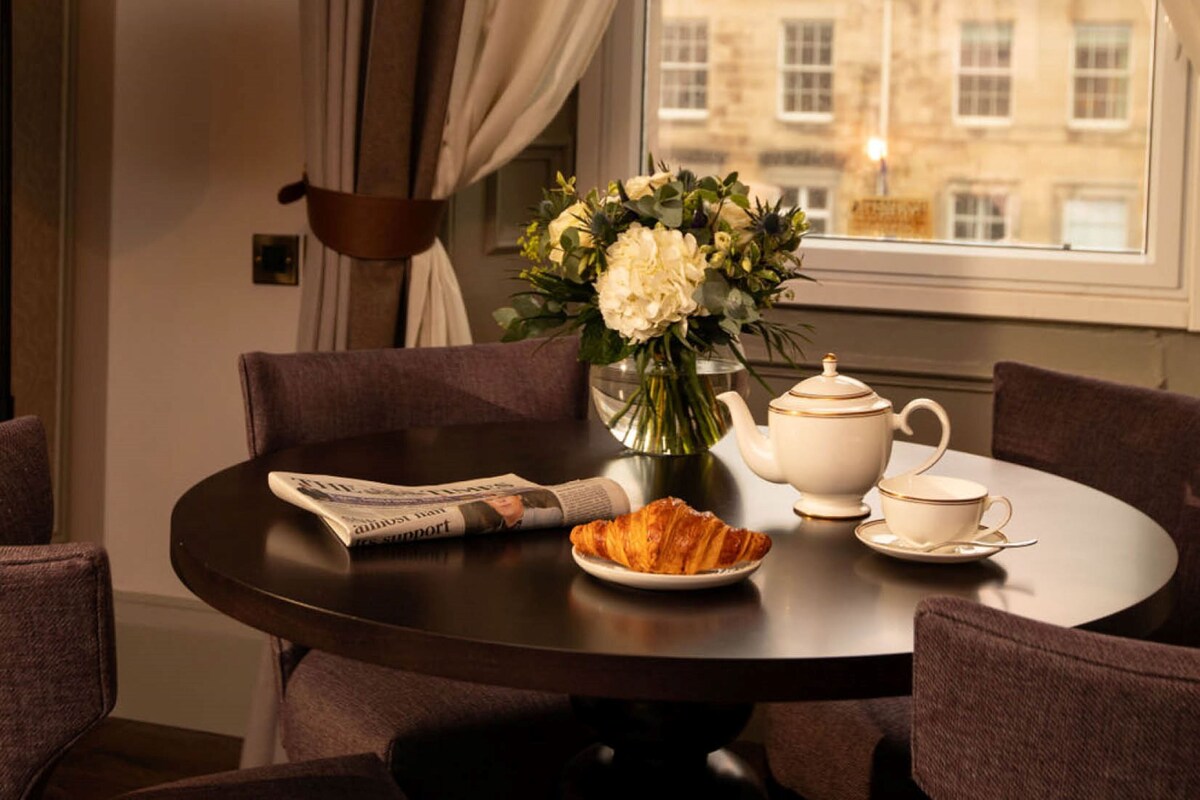 A round table with a newspaper, a croissant, a teapot, and a cup set. There is a vase with flowers on the table.