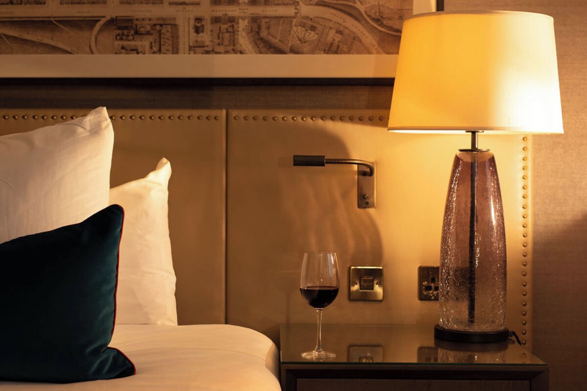 A hotel room with a bed, pillows, a bedside table with a glass of red wine, a lamp, and a wall-mounted phone.
