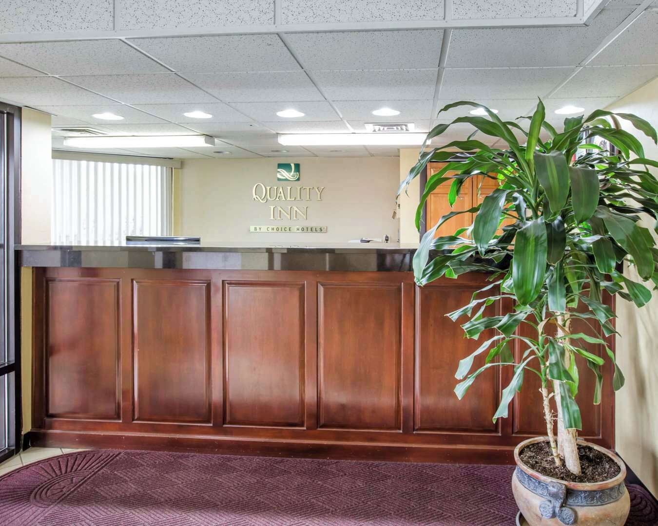 A reception area with a wooden reception desk, a potted plant, and a sign for 'Quality Inn by Choice Hotels'.