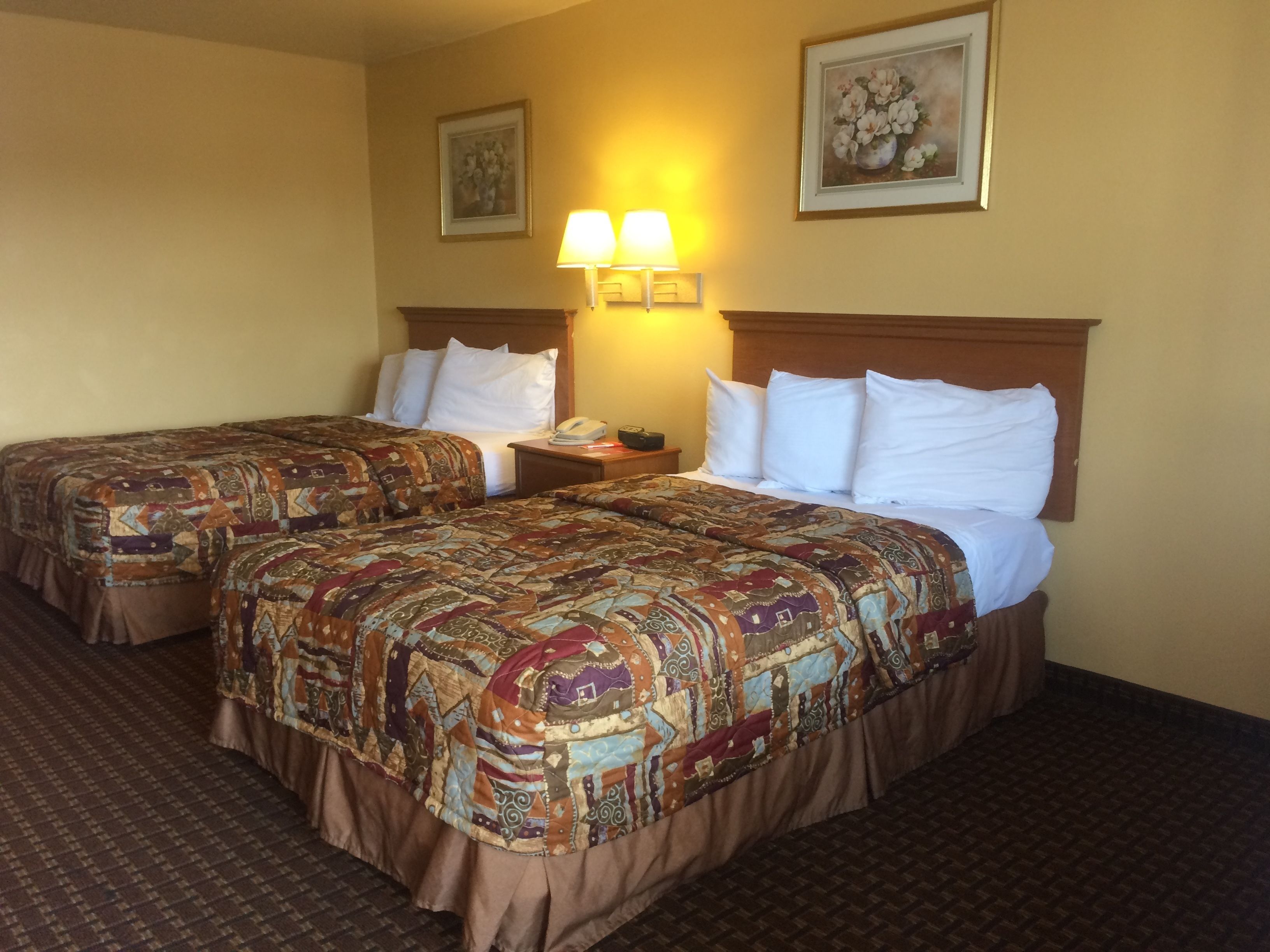 A hotel room with two beds, each with a patterned bedspread and white pillows. There is a nightstand with a lamp and a telephone. The room has a carpeted floor and a painting on the wall.