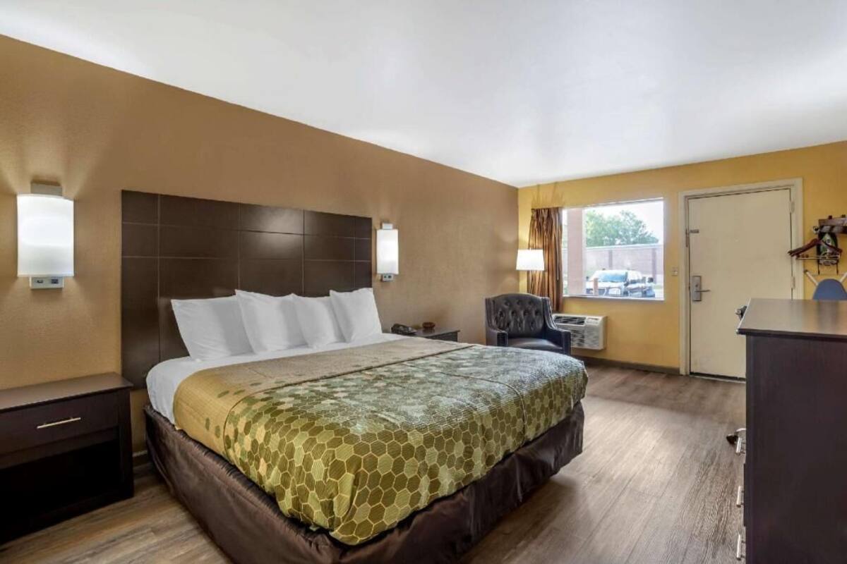 Ein Hotelzimmer mit einem großen Bett, einem Nachttisch, einem Sessel, einem Schreibtisch und einem Fenster mit Blick ins Freie.
