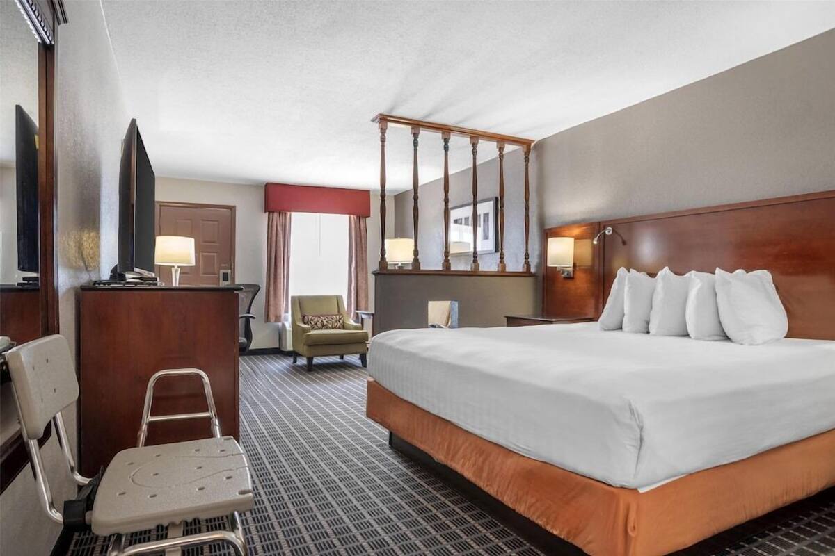 Une chambre d’hôtel avec un grand lit, un bureau, une chaise, un fauteuil roulant, un miroir et une fenêtre avec des rideaux.