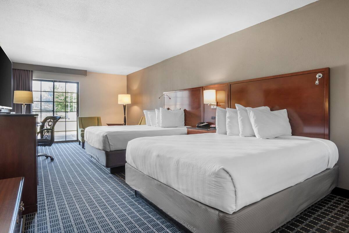A hotel room with two beds, a desk, a chair, a lamp, and a window.