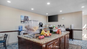 Un espace pour le petit-déjeuner à l’hôtel, avec un comptoir en granit, une machine à café, un panier de fruits et une horloge au mur.