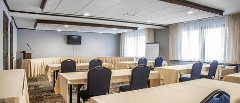 A conference room with tables and chairs, a podium, a screen, and large windows with curtains.