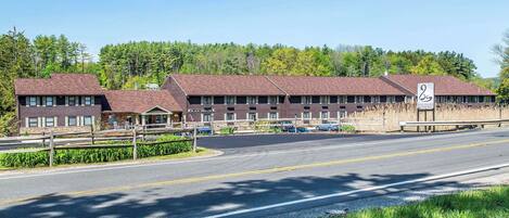 A motel with a central building and two wings, a paved road in front, and a forested backdrop.