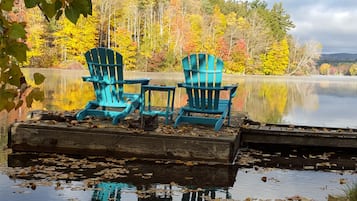 Dos sillas Adirondack azules sobre un muelle, frente a un lago tranquilo, rodeado de follaje otoñal.