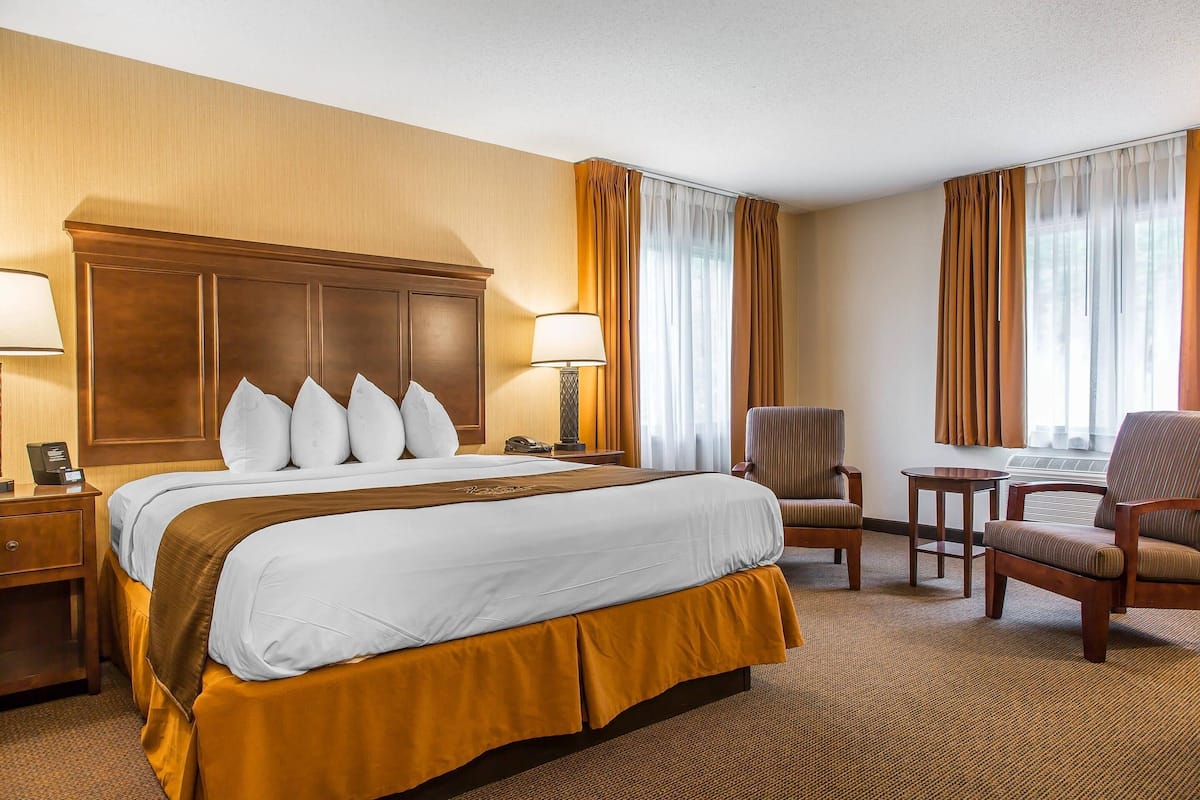 Ein Hotelzimmer mit einem großen Bett, zwei Nachttischen, einem Sessel, einem kleinen Tisch und einem Fenster mit Vorhängen.