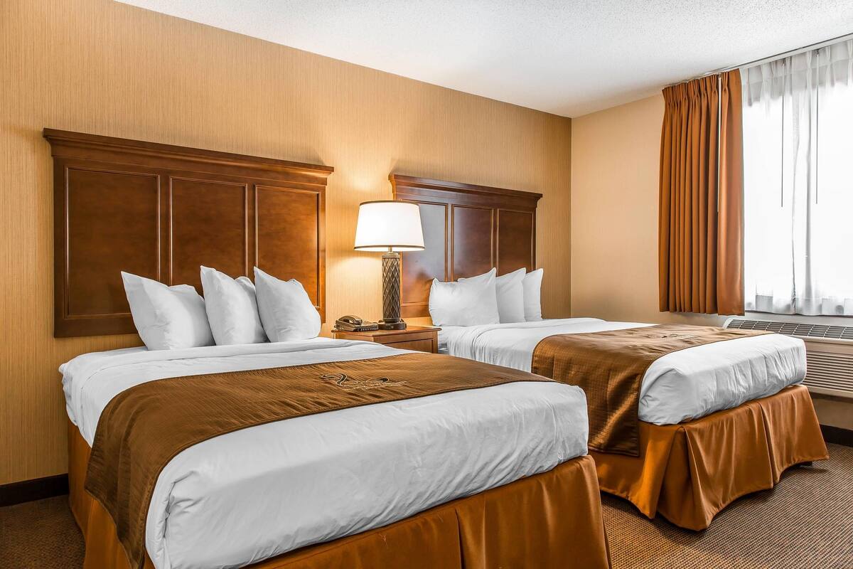Ein Hotelzimmer mit zwei Betten, Holz-Kopfteilen, einem Nachttisch mit Lampe und einem großen Fenster mit Vorhängen.