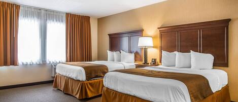 A hotel room with two beds, a large wooden headboard, a bedside table with a lamp, and a large window with curtains.