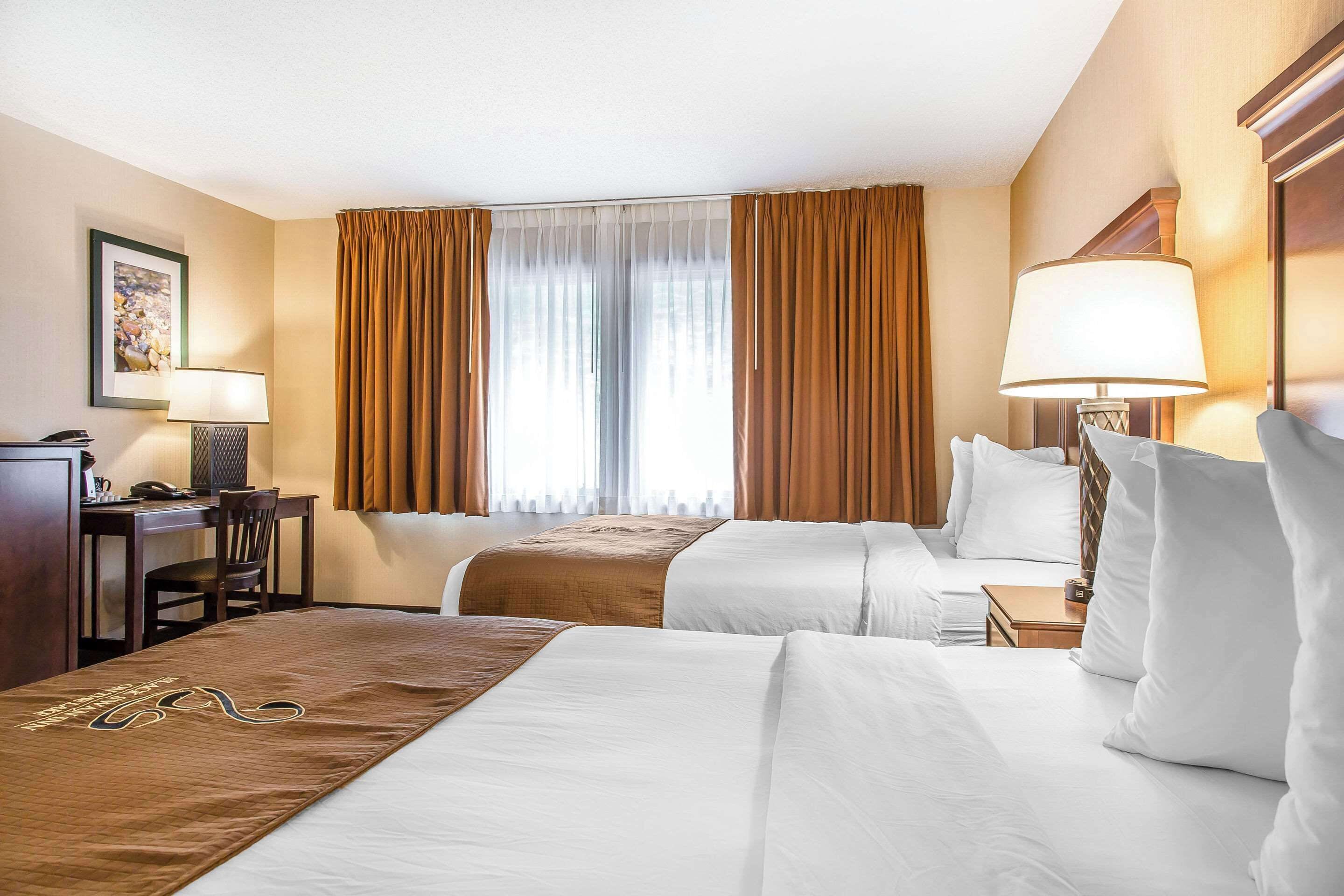 A hotel room with two beds, a desk, a chair, and a lamp.