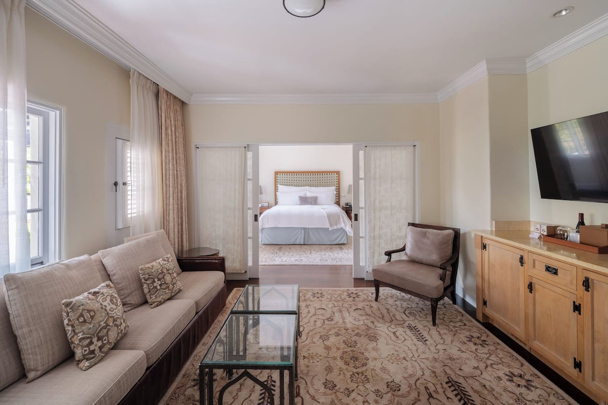 A hotel room with a sofa, armchair, glass coffee table, wooden cabinet, and a television.