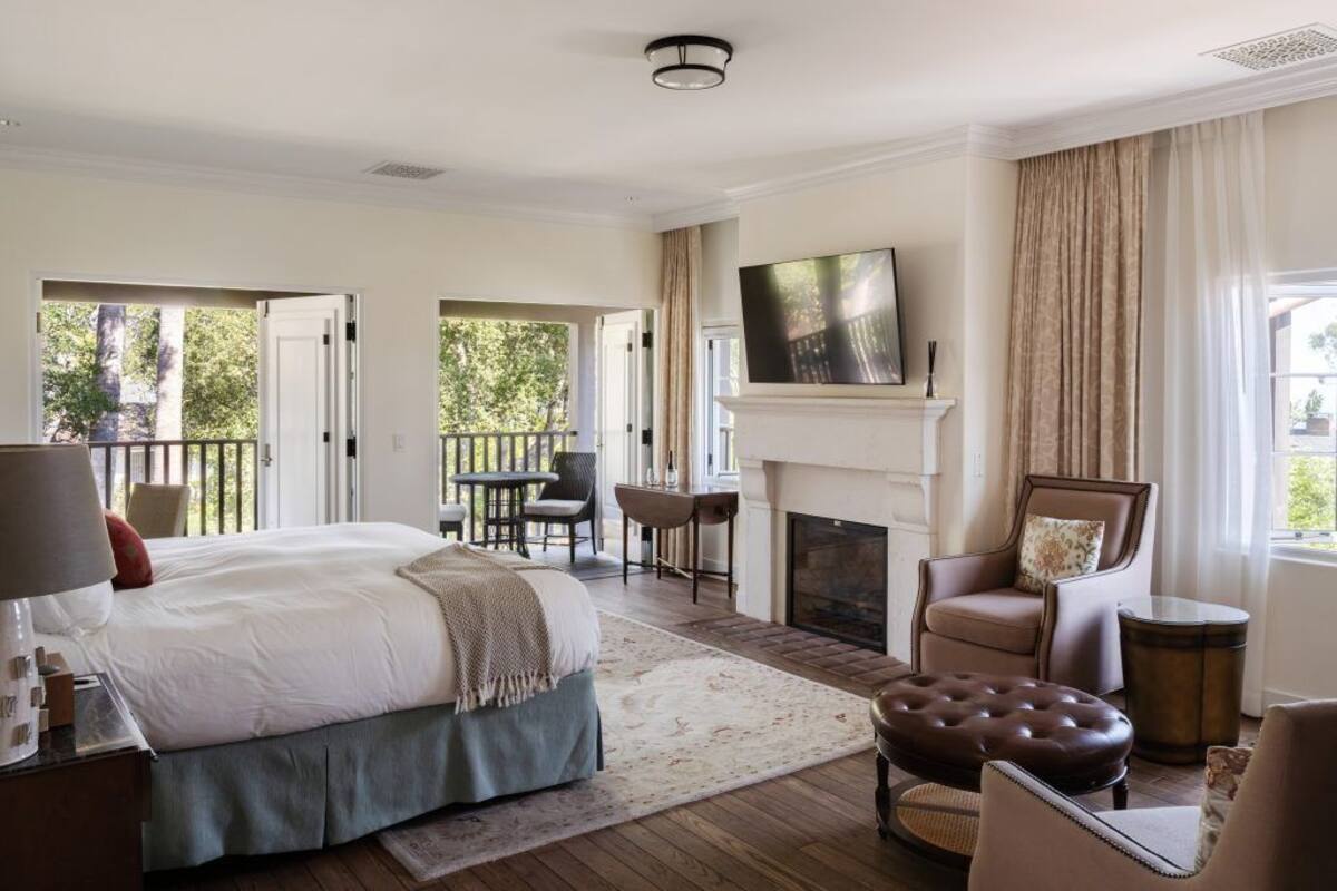A spacious bedroom with a large bed, a fireplace, a balcony, and a view of greenery.