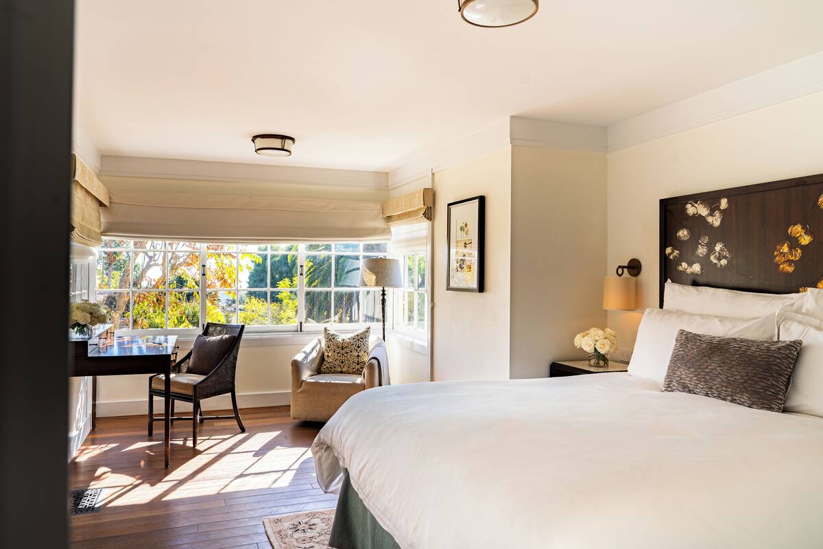 A bedroom with a large bed, a desk, a chair, and a window with a view of trees.