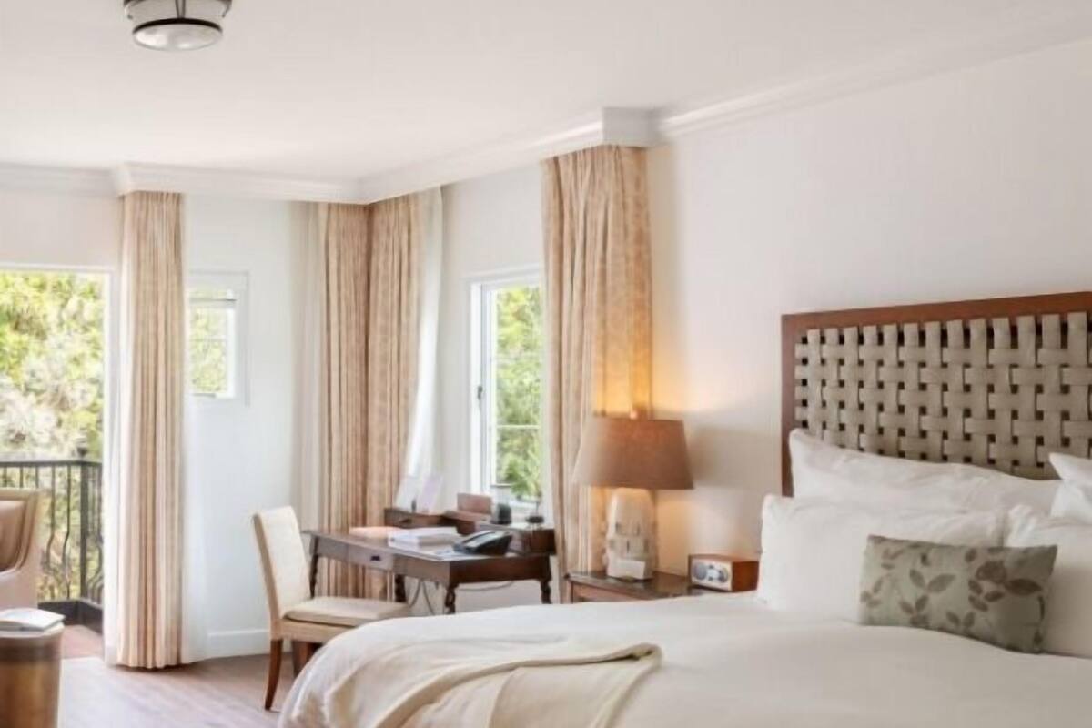 A hotel room with a large bed, a desk, a chair, a lamp, and a view of greenery outside.