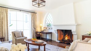 A living room with a fireplace, a sofa, a chair, a table with books and a vase of flowers, and a window with curtains. - El Encanto Santa Barbara (Santa Barbara)