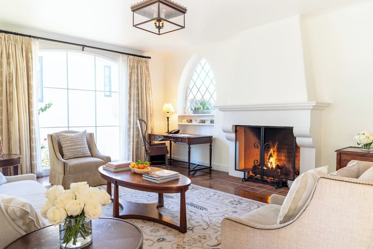 A living room with a fireplace, a sofa, a chair, a table with books and a vase of flowers, and a window with curtains.