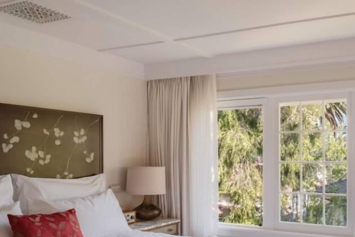 A hotel room with a bed, a bedside table with a lamp, a window with a view of greenery, and a decorative headboard.