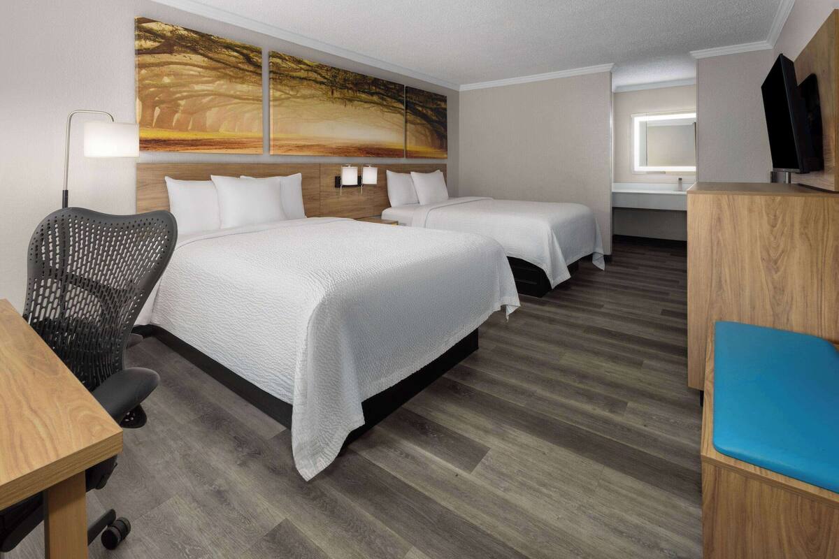 A hotel room with two beds, a desk, a chair, a TV, and artwork on the wall.