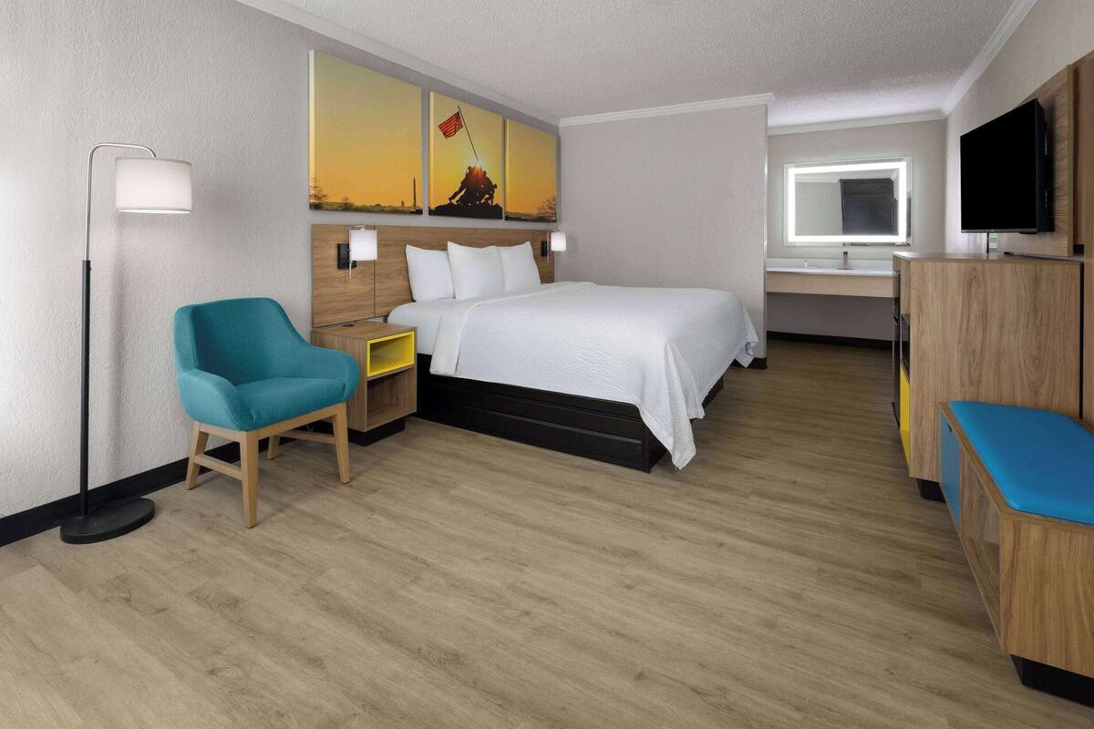 A hotel room with a bed, a chair, a desk, a TV, and a painting on the wall.