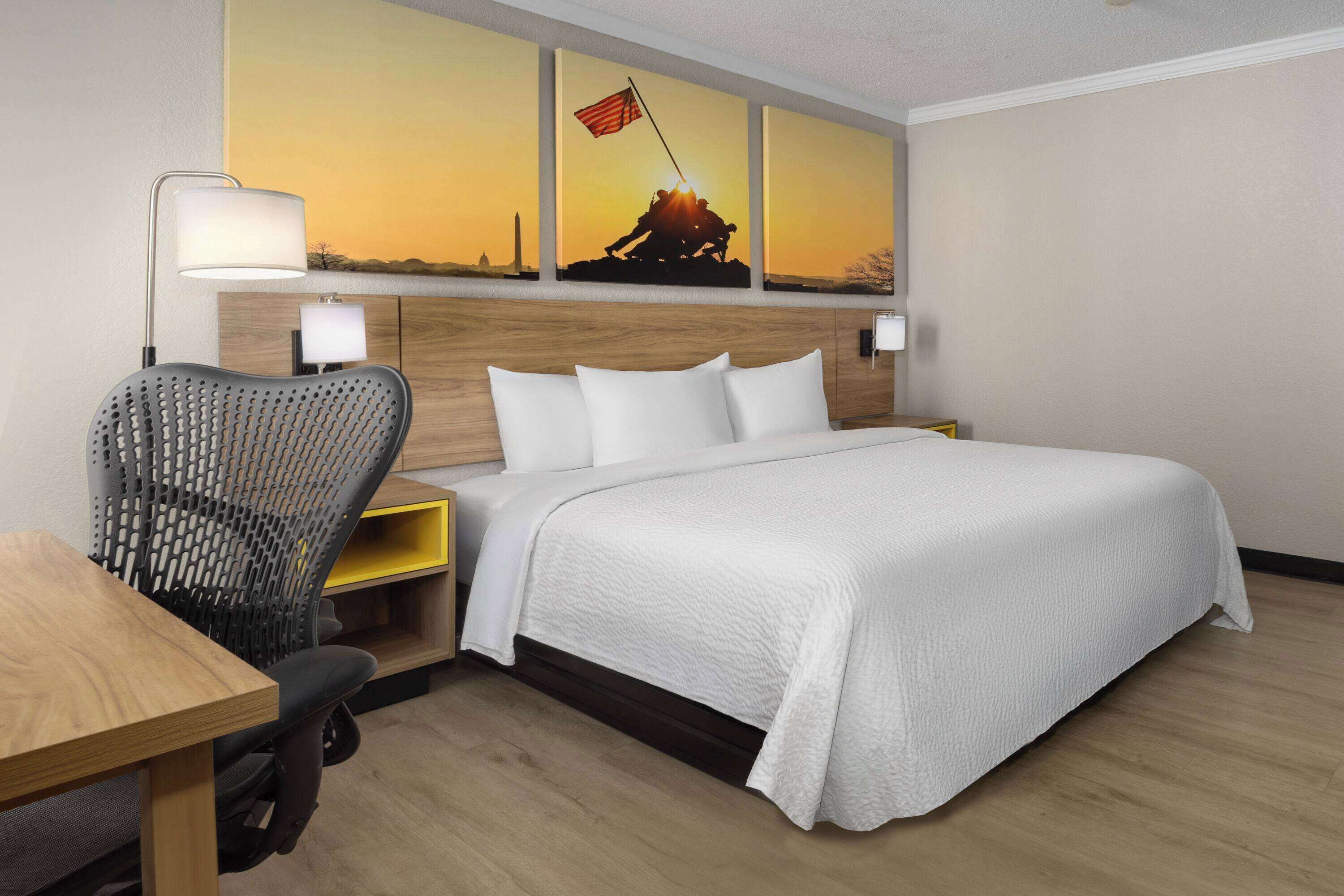 A hotel room with a bed, a desk, a chair, and a wall-mounted lamp.