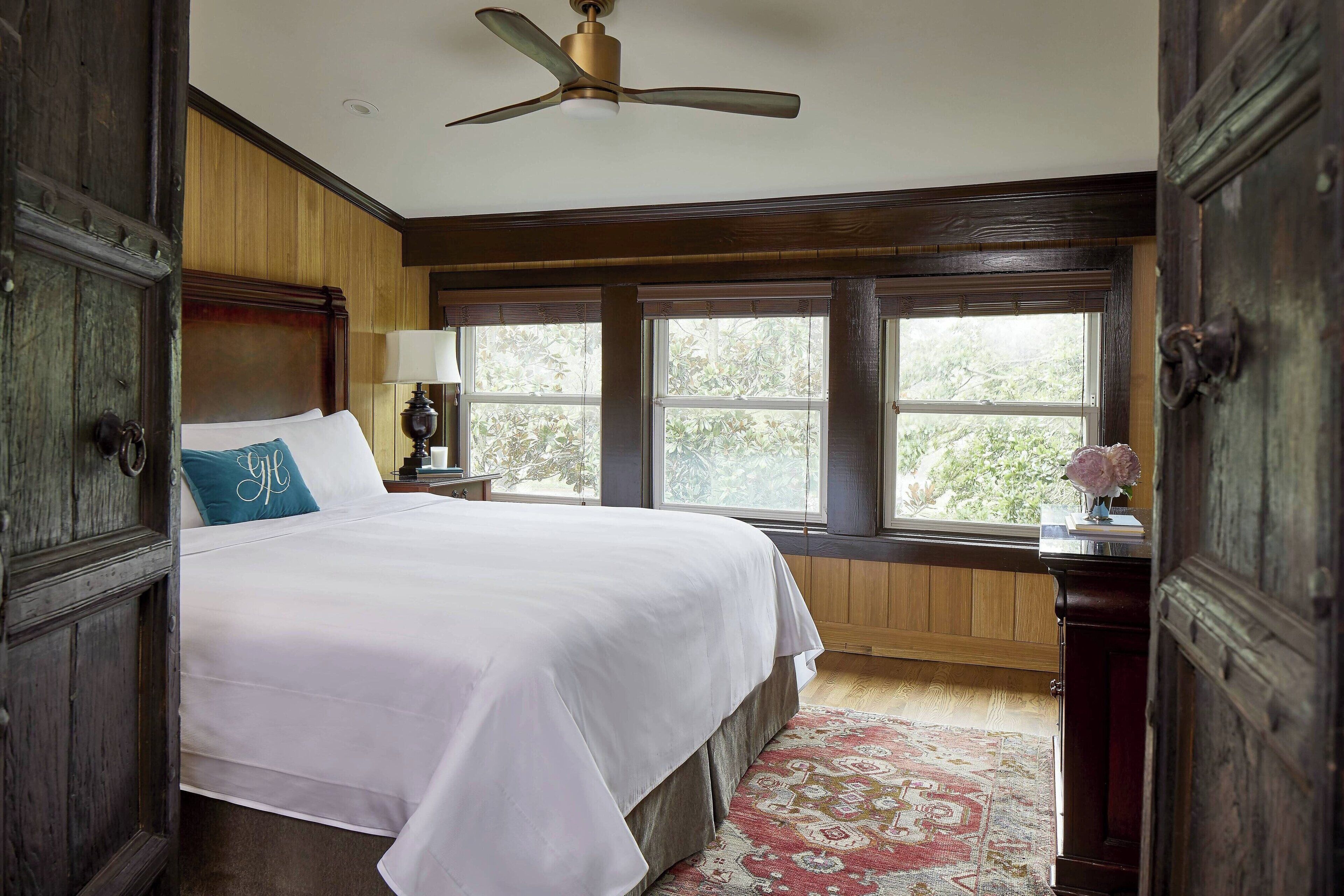 A bedroom with a large bed, wooden walls, a ceiling fan, and a view through the windows.