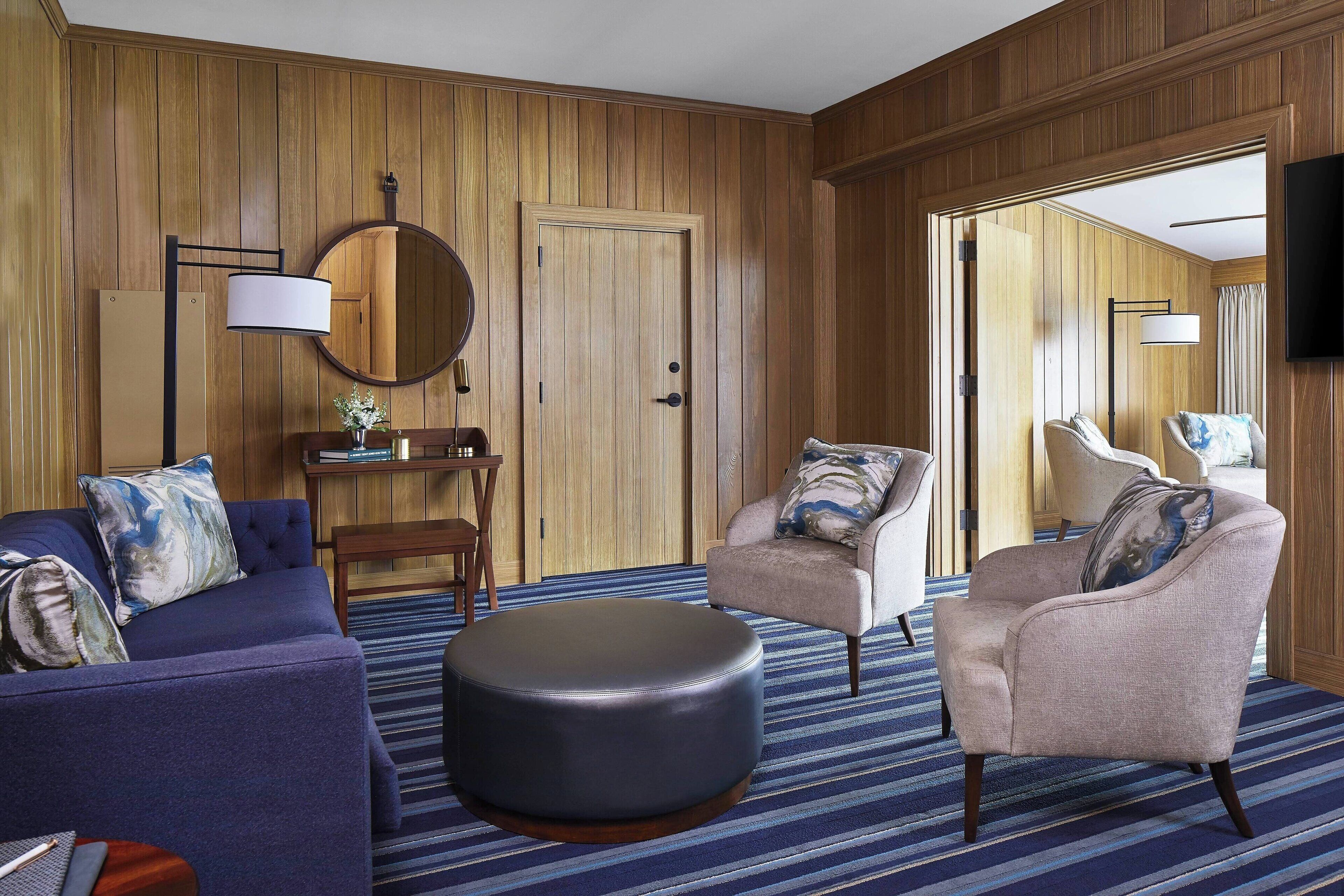 A room with a blue sofa, a round ottoman, a beige armchair, and a wooden door.