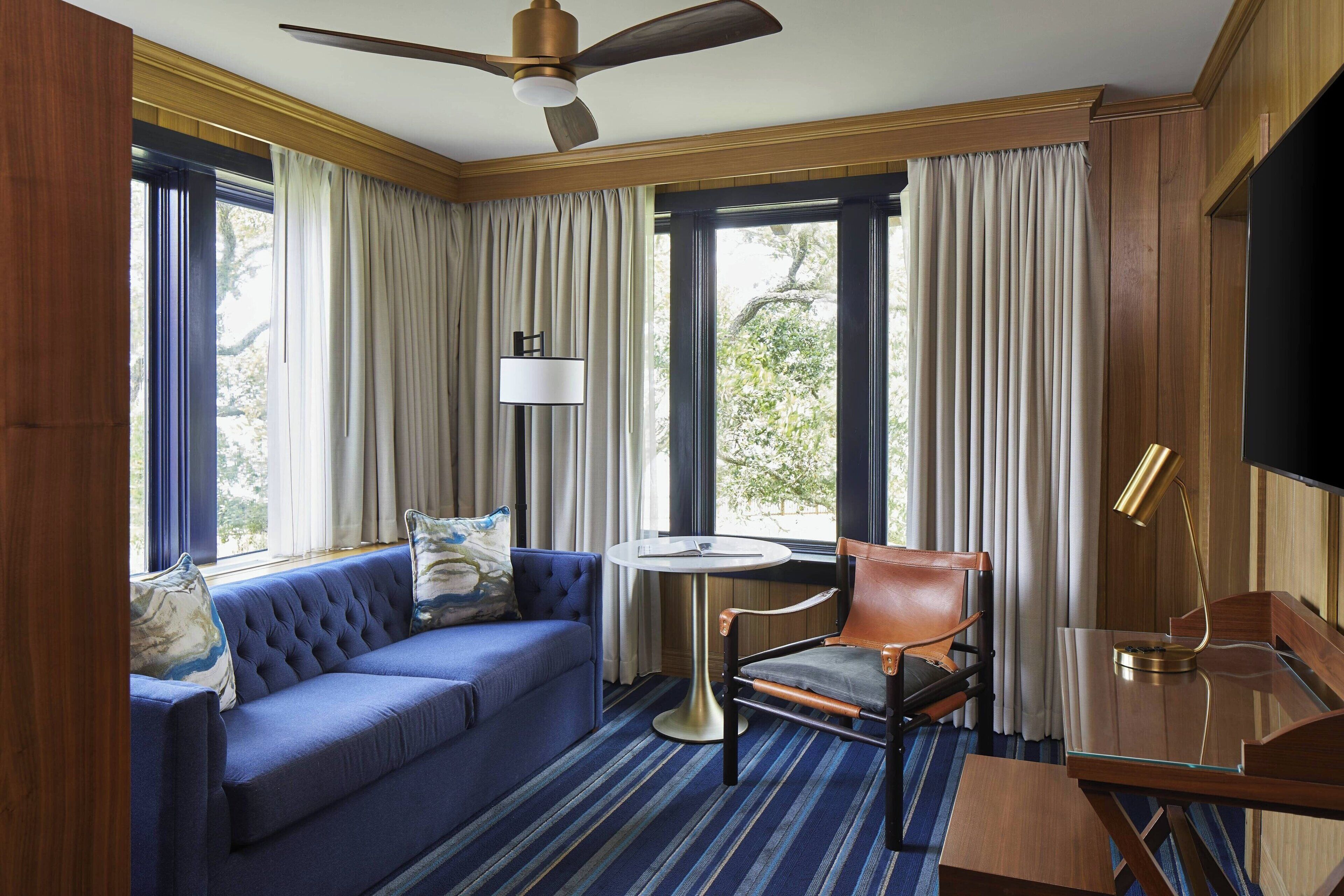 A room with a blue sofa, a wooden chair, a small round table, a ceiling fan, and large windows with curtains.