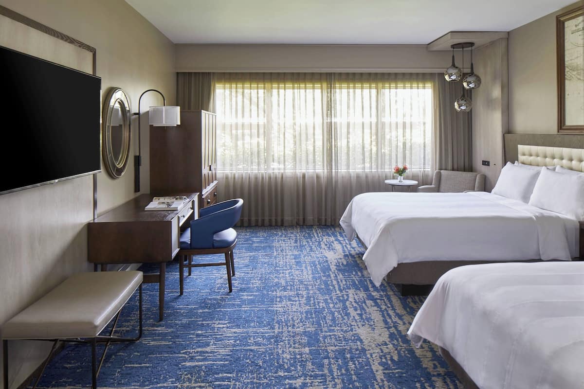 A hotel room with two beds, a desk with a chair, a TV, and a large window with curtains.