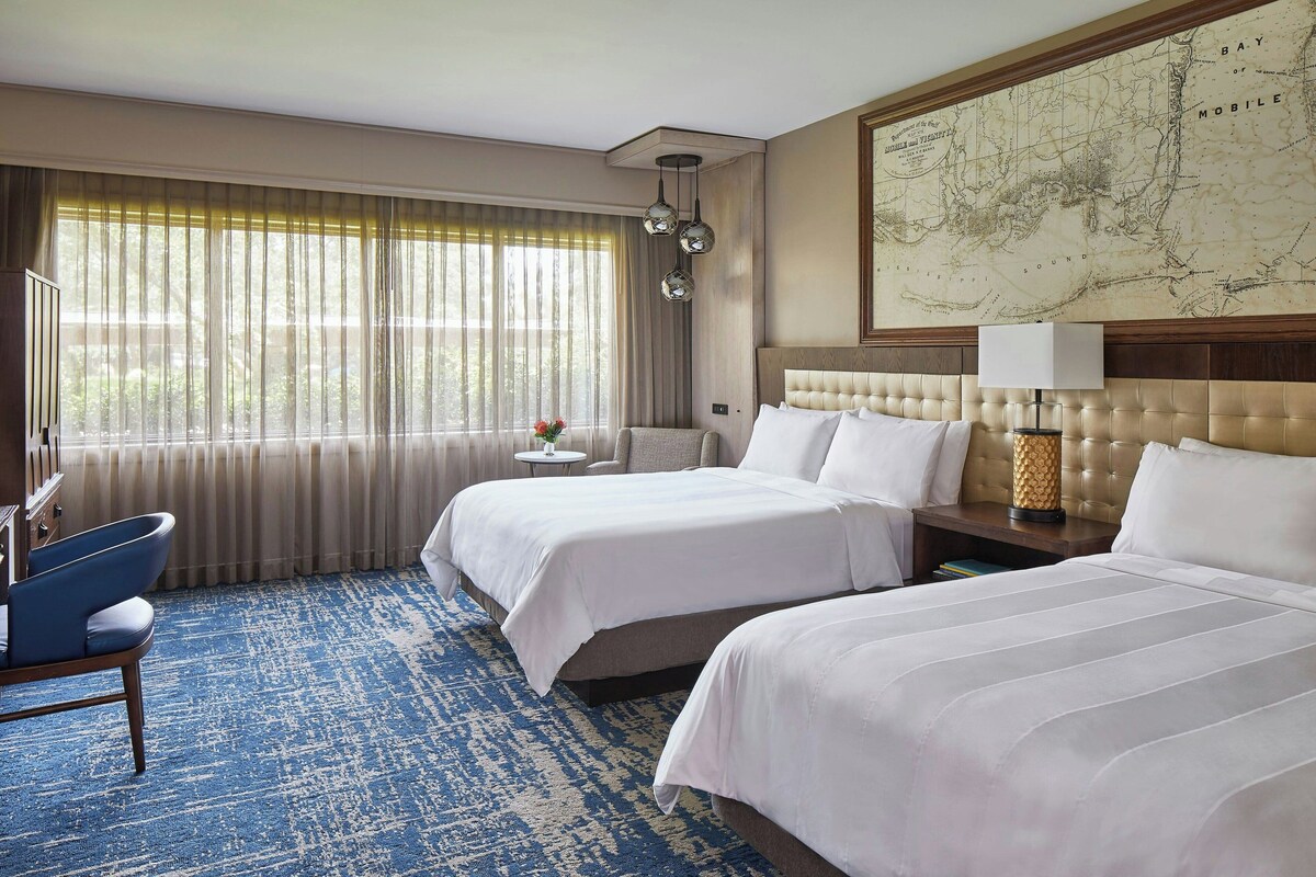 A hotel room with two beds, a large map on the wall, a blue carpet, and a view of the outdoors through the window.