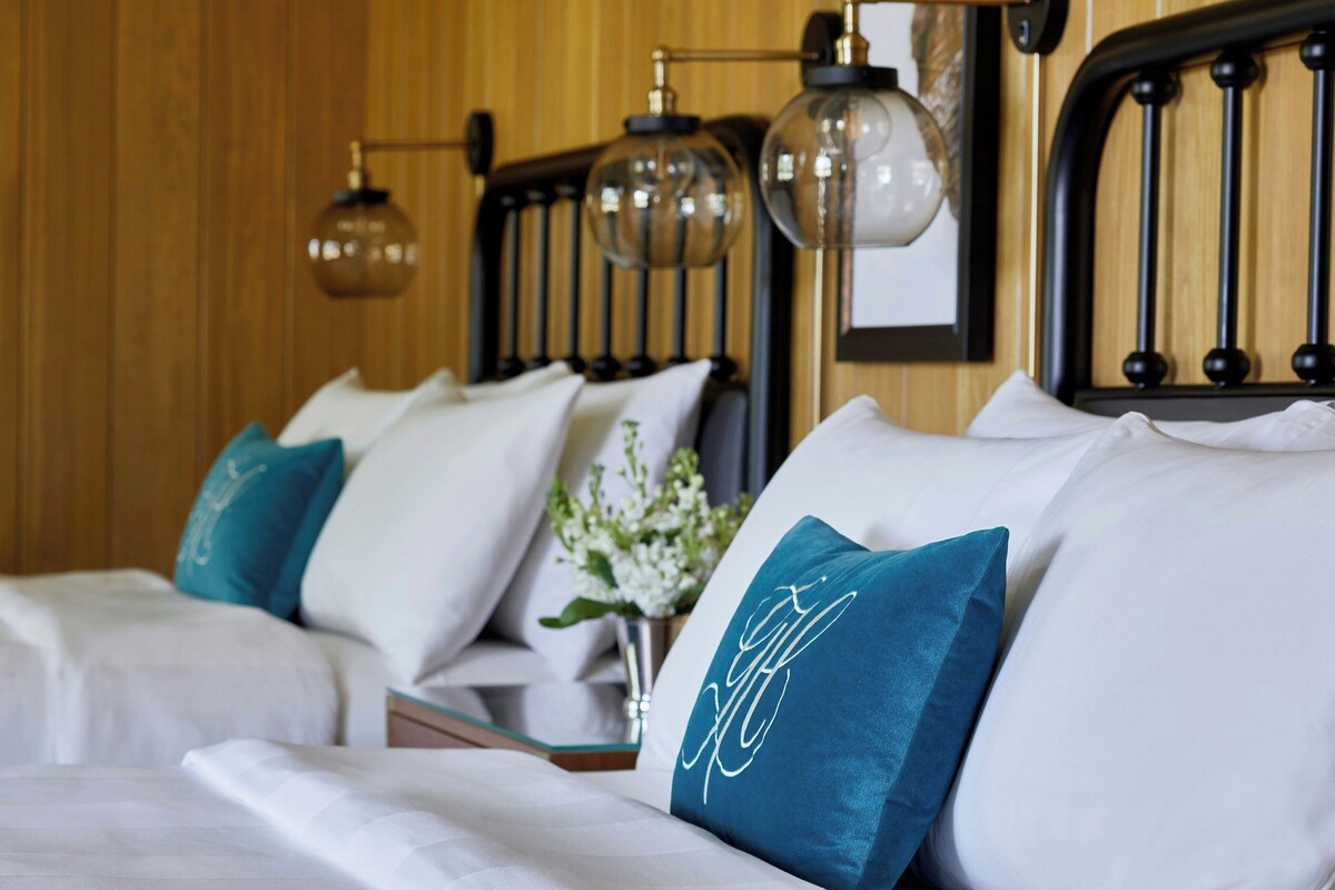 A hotel room with two beds, each with white bedding and teal pillows, a wooden headboard, and hanging spherical lamps.