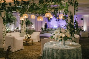 Salón de recepción de bodas con mobiliario blanco, arreglos florales e iluminación colgante.