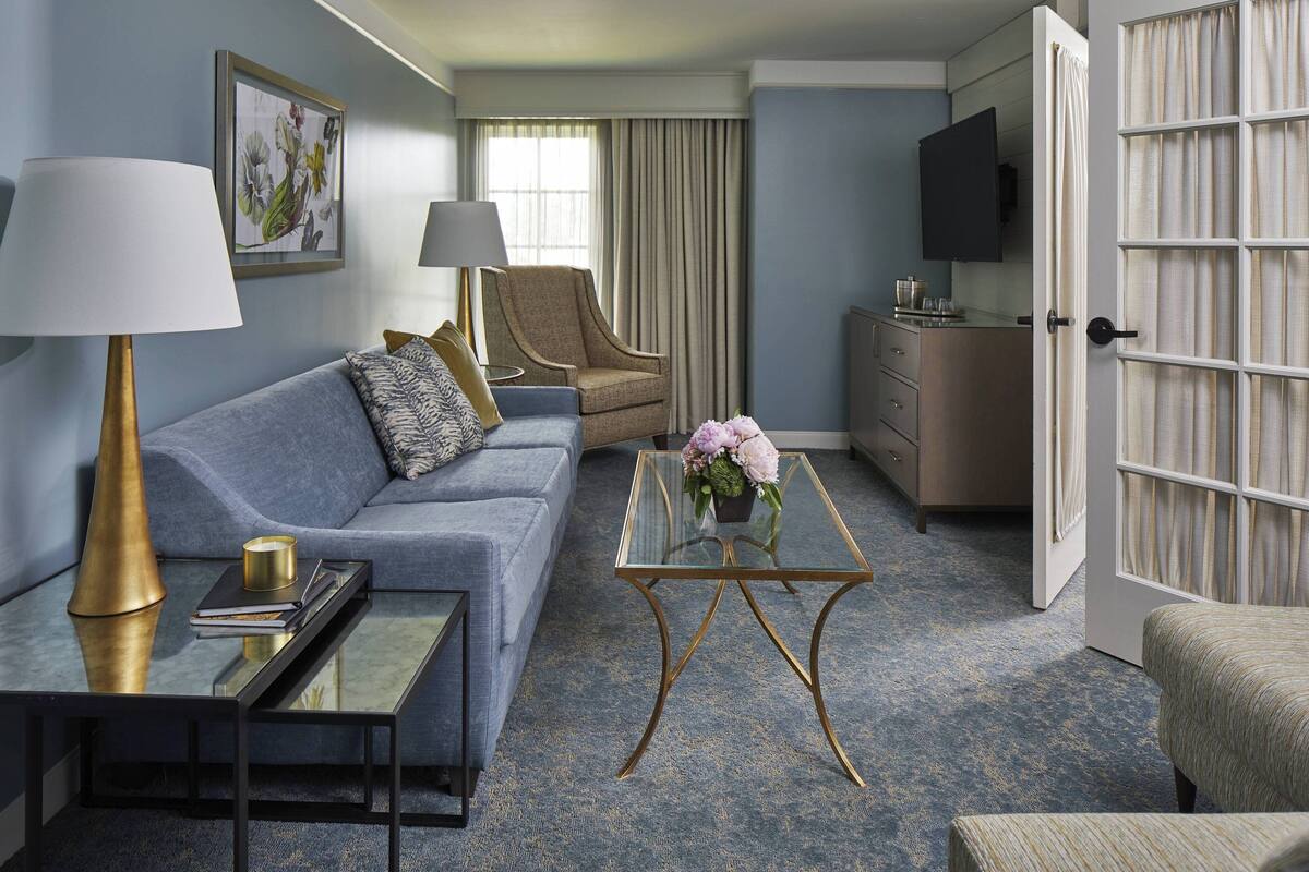 Une chambre d’hôtel avec un canapé bleu, une table basse en verre, un fauteuil beige, une télévision et un mini-bar.