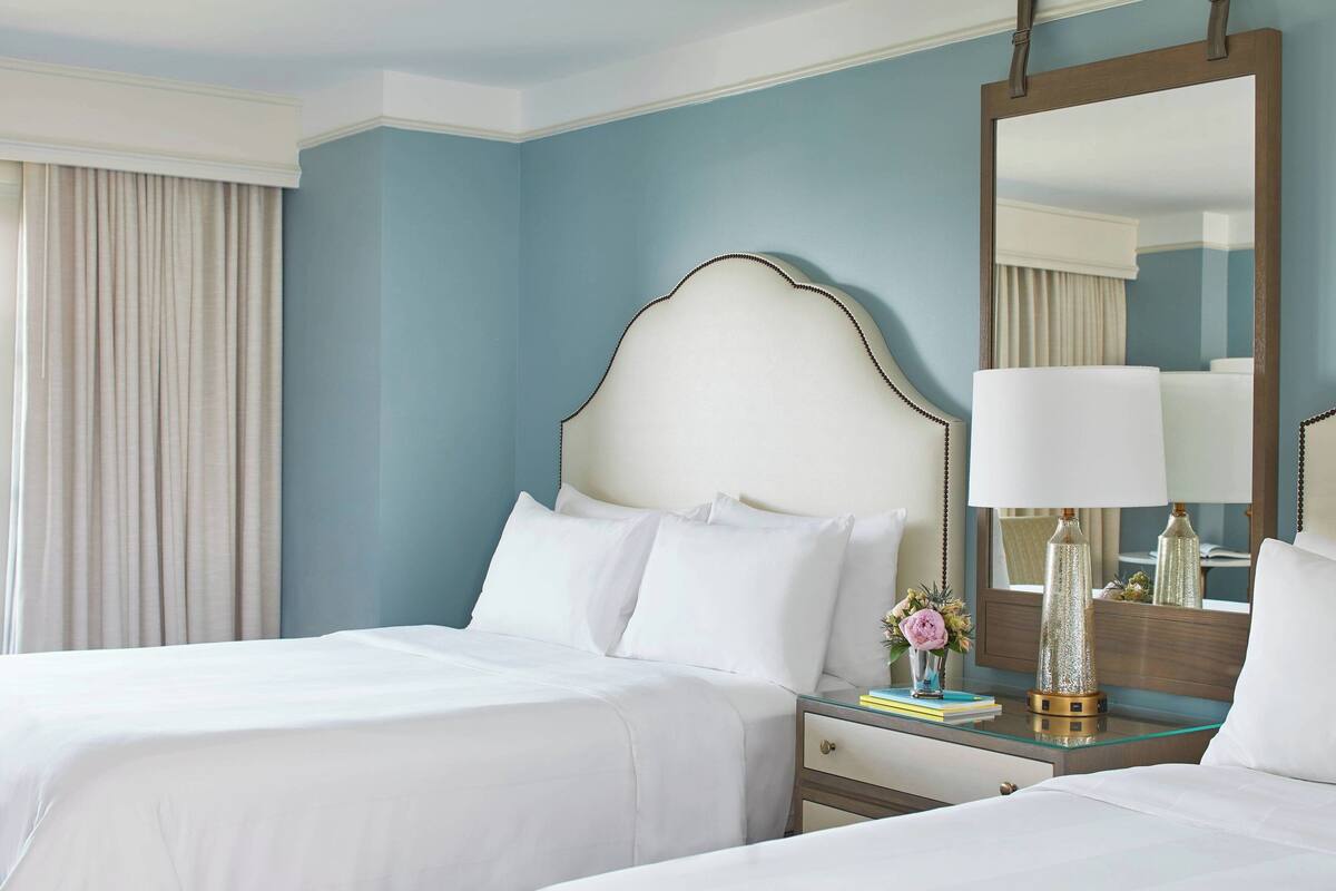 Une chambre d’hôtel avec deux lits, un grand miroir, une table de chevet avec une lampe et une fenêtre avec des rideaux.