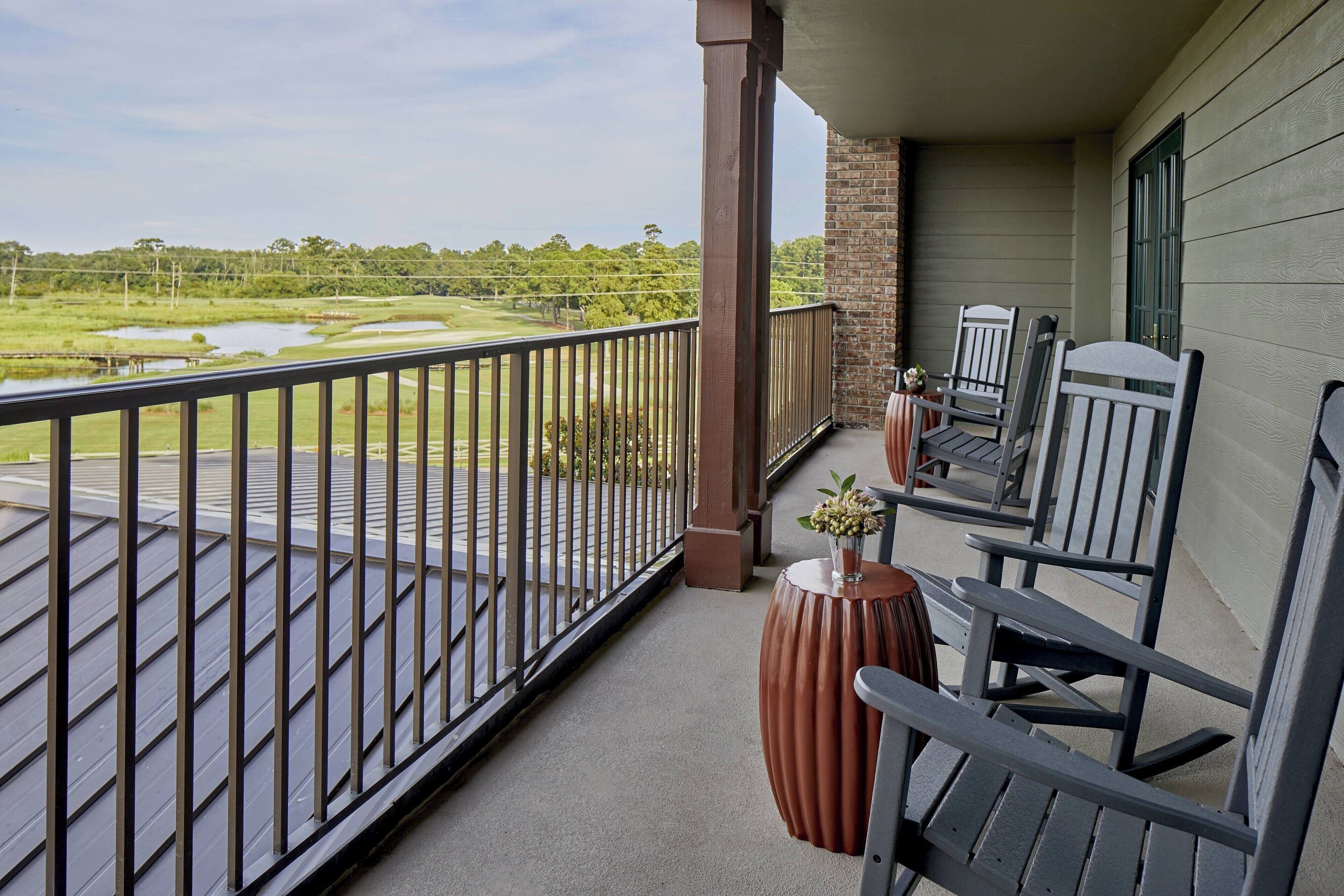 A balcony with a view of a golf course, a wooden railing, and two rocking chairs.