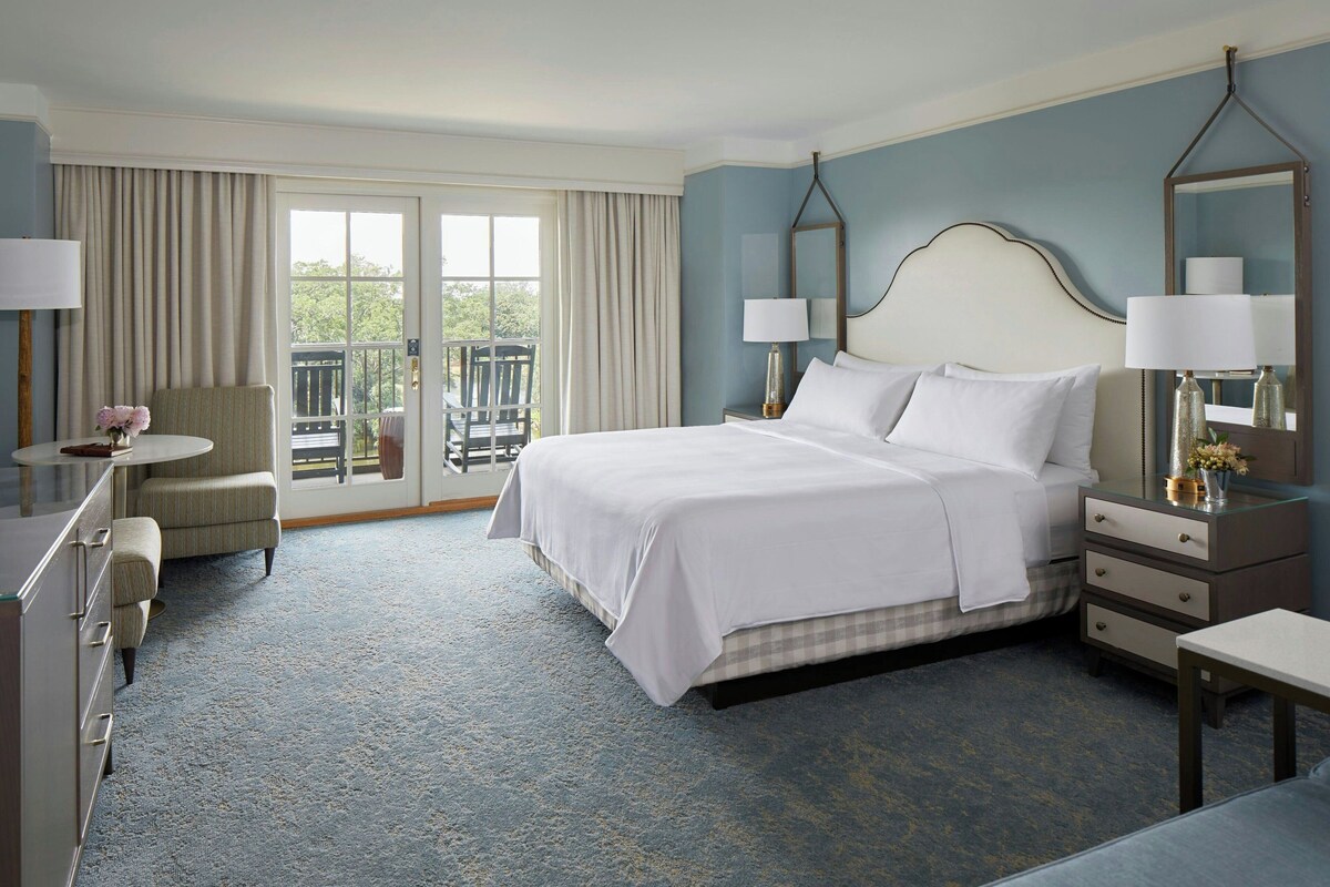 A hotel room with a bed, a chair, a desk, a dresser, and a balcony with a view.