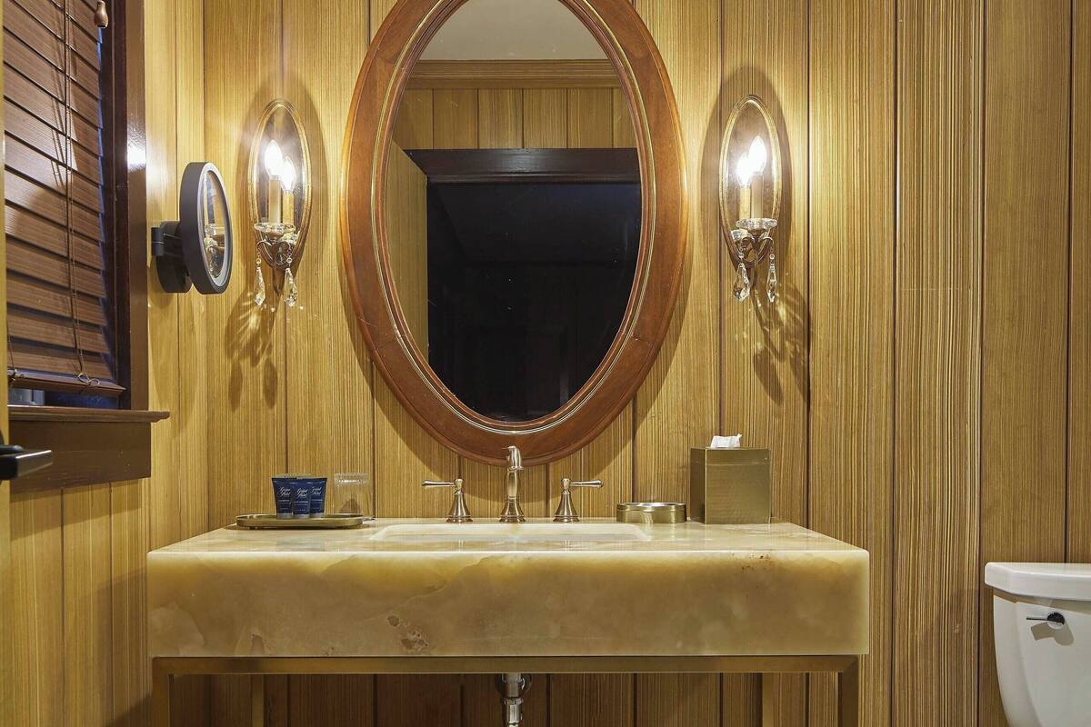 Une salle de bain avec un lambris en bois, un miroir rond, un lavabo avec un plan de toilette en marbre et des toilettes.