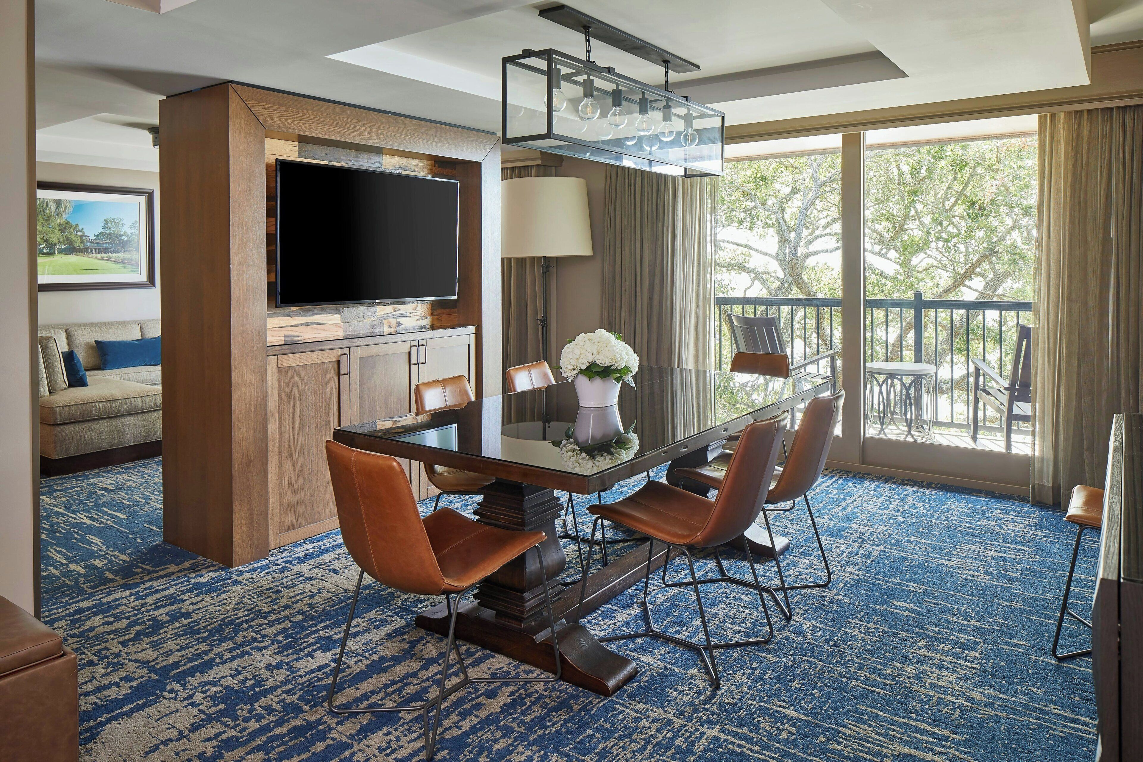 A hotel room with a large flat-screen TV, a dining table with chairs, a sofa, a balcony with outdoor furniture, and a large window.