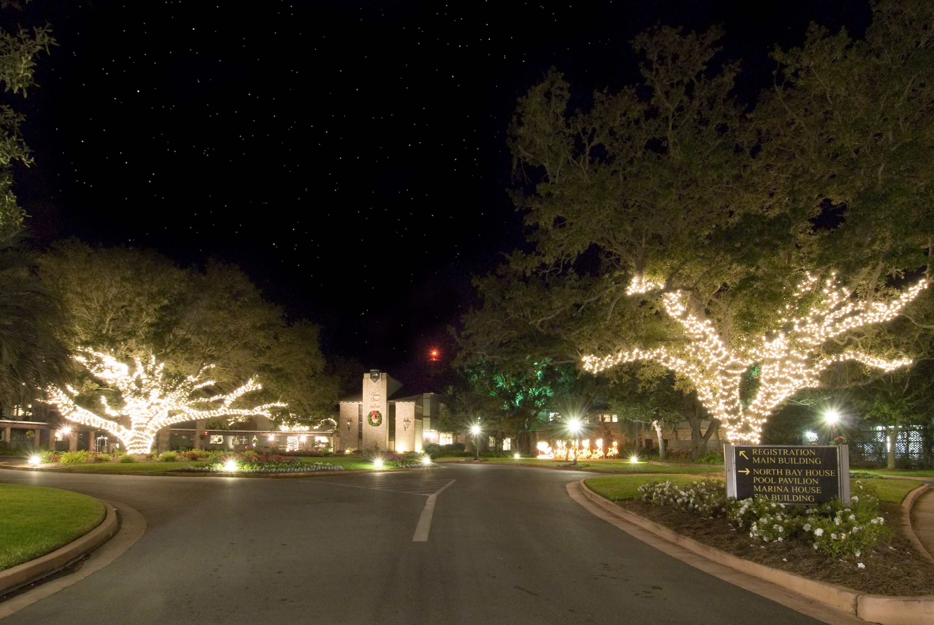 Vista noturna de um campus com árvores iluminadas e uma placa indicando direções para vários edifícios.