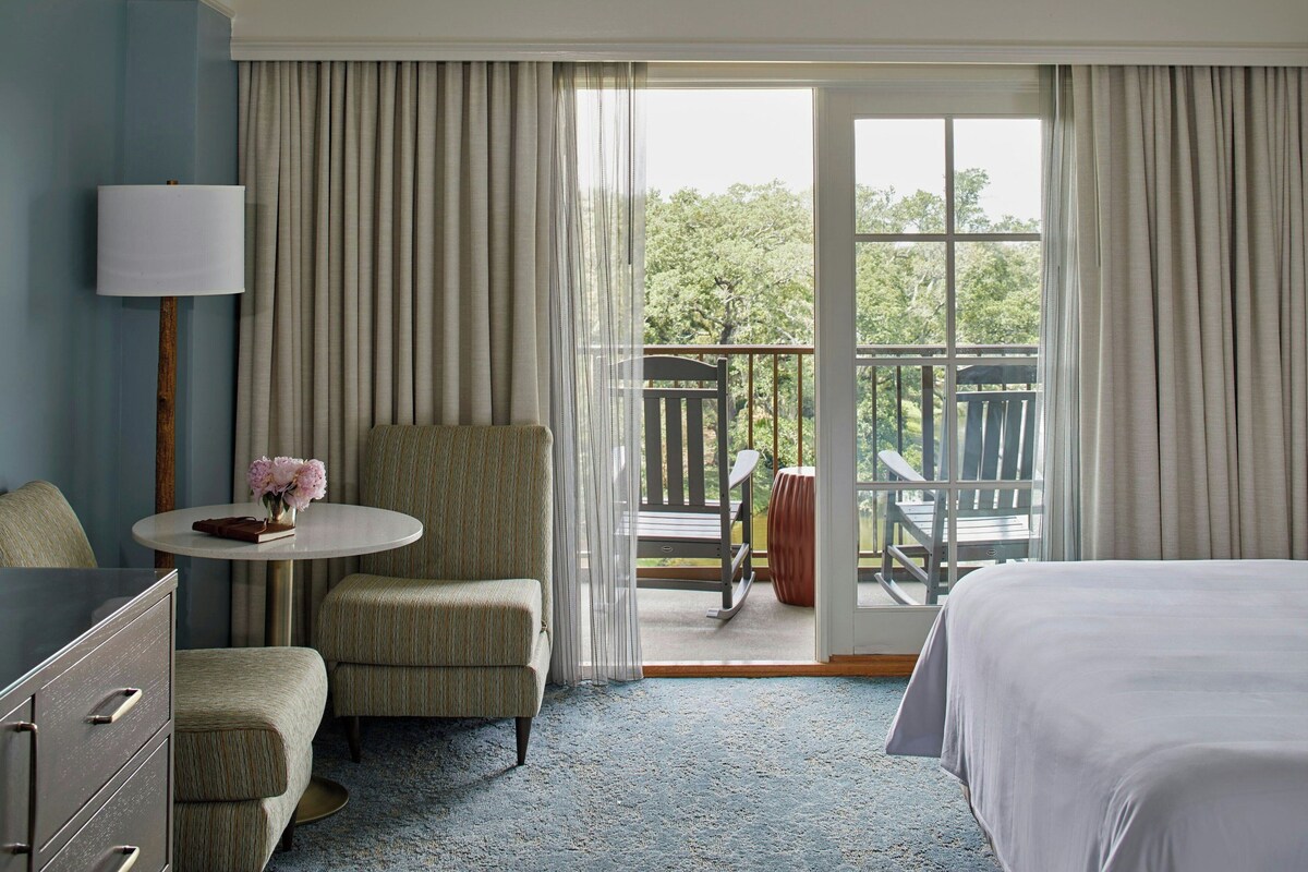 A hotel room with a bed, a chair, a small table with a vase, a lamp, and a view of the outdoors through a sliding glass door.