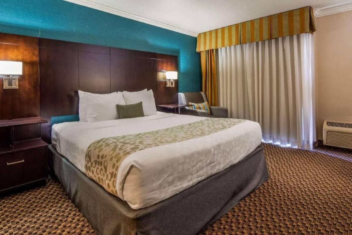 A hotel room with a large bed, a nightstand, a desk, a chair, a window with curtains, and an air conditioning unit.